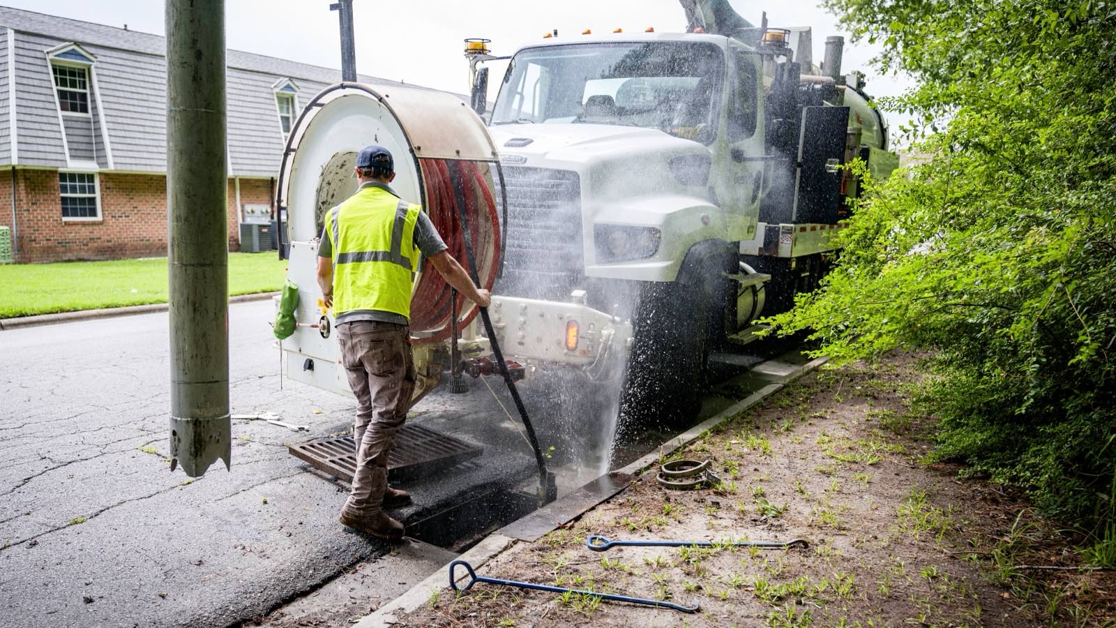 Worker in a hi-vis vest at a street drain beside a vacuum truck, with water spraying