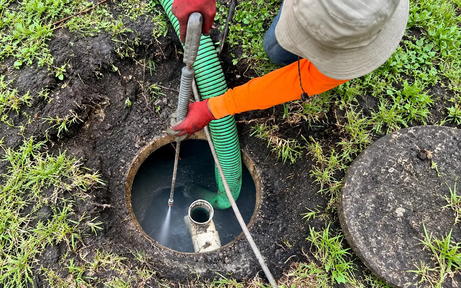 Worker spraying a pressure wand into an open septic tank while a vacuum hose drains it