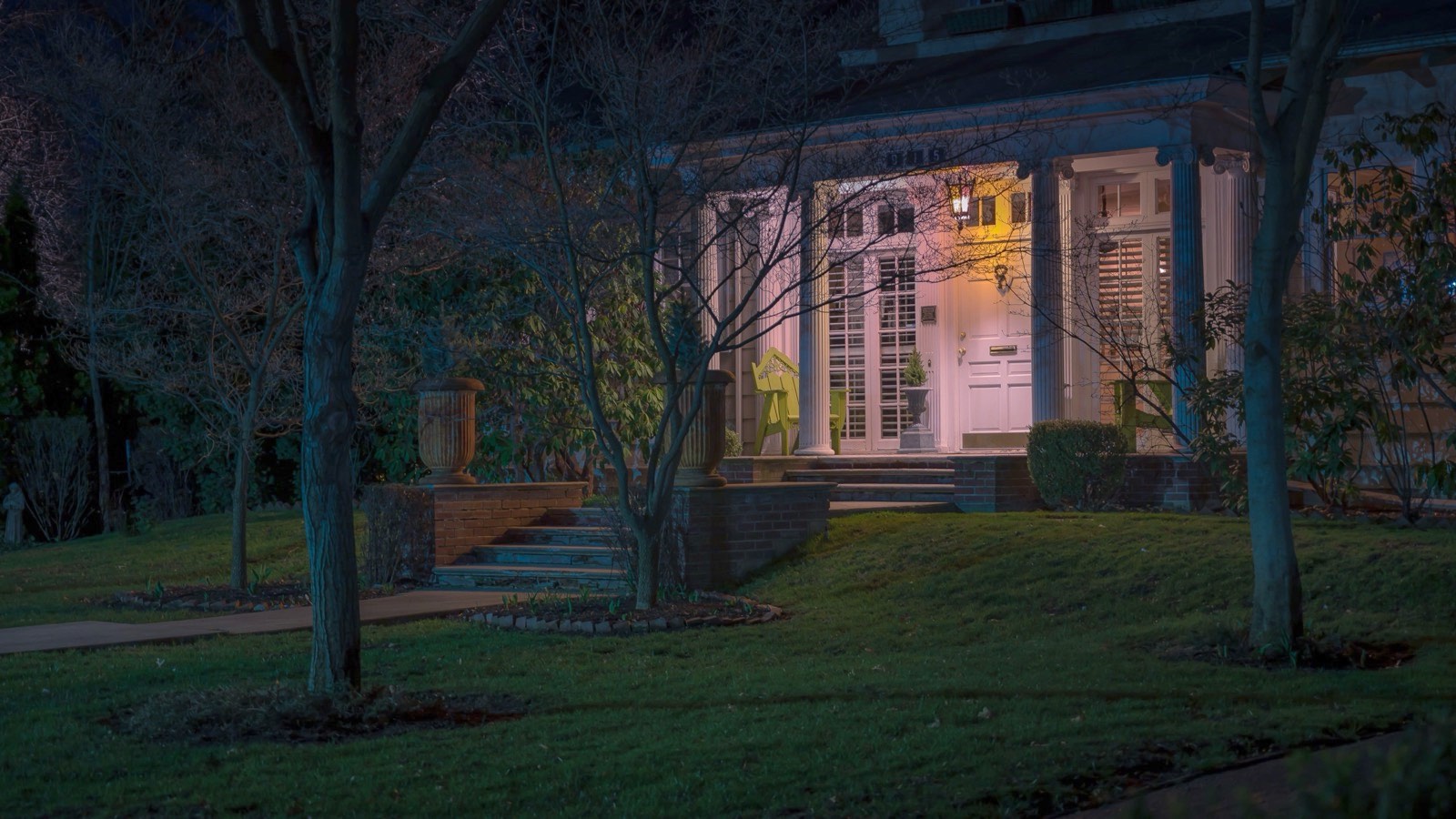Front porch of a house lit up at night behind a lawn and bare trees