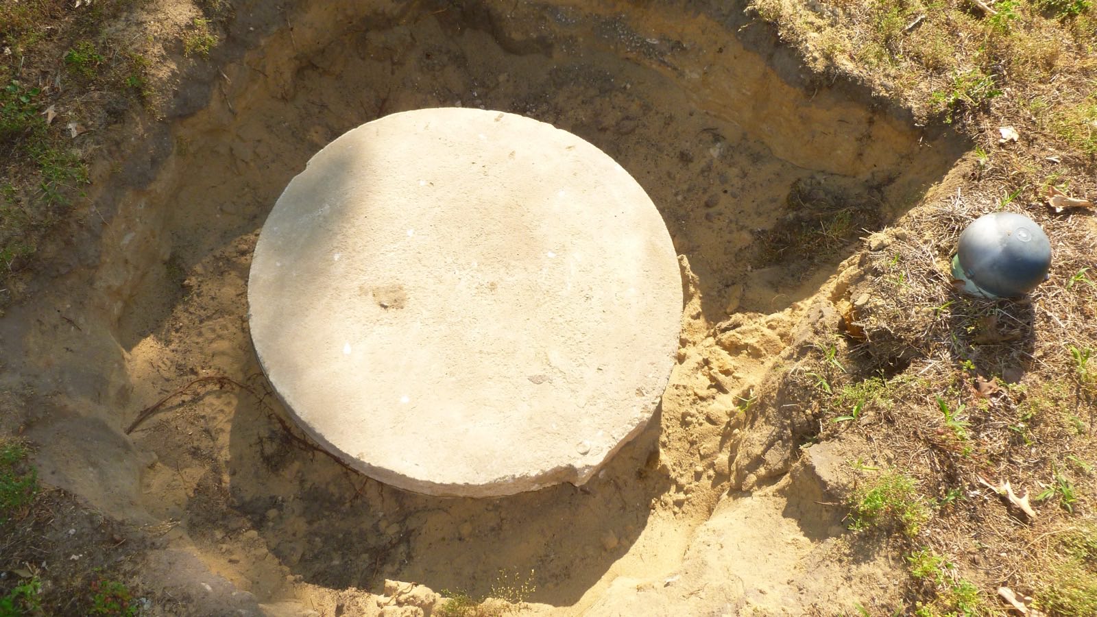 Round concrete lid sitting in a sandy hole in the ground, seen from above