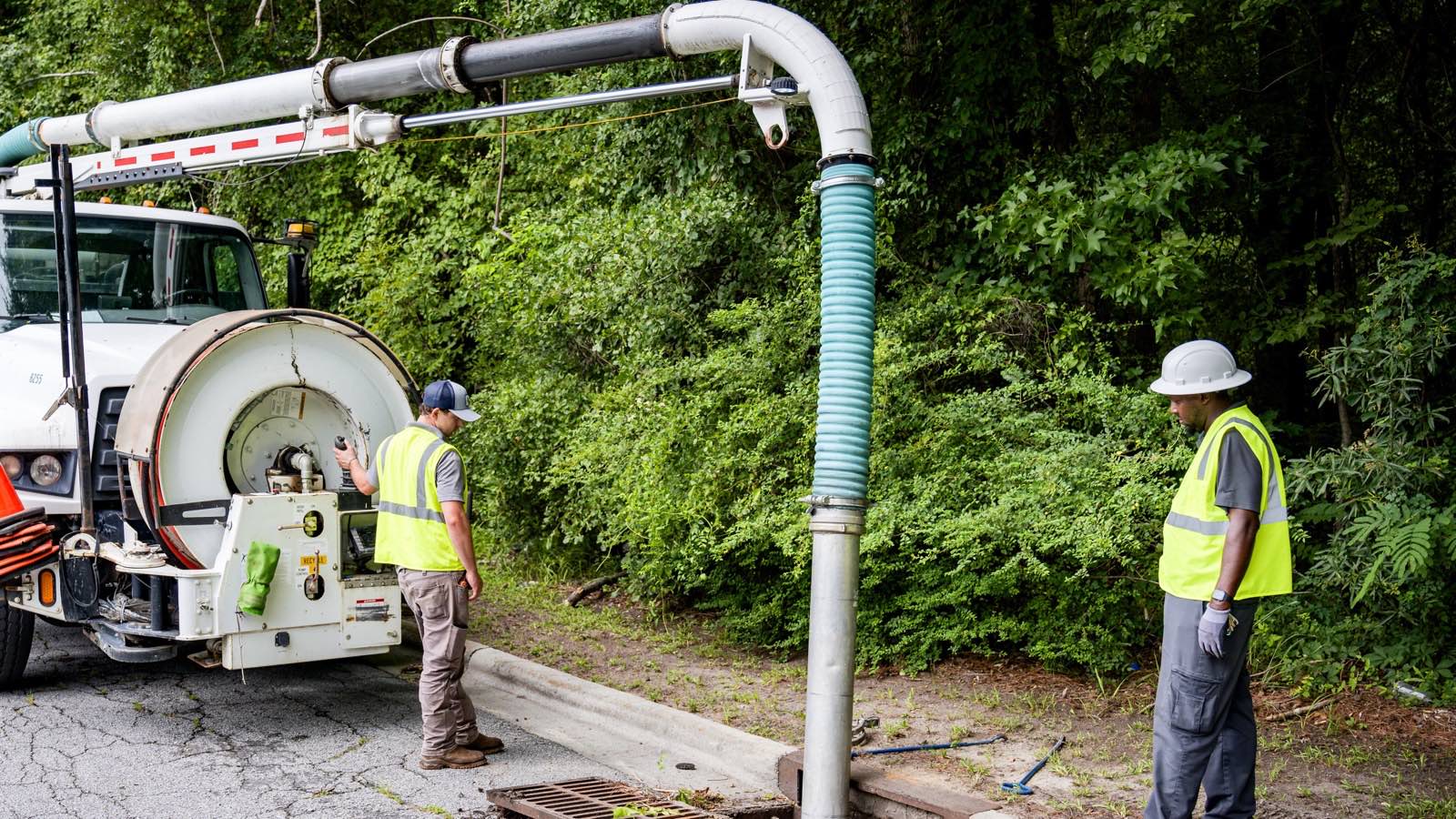 Vacuum truck with a boom and green hose beside a road, with two workers in hi-vis vests